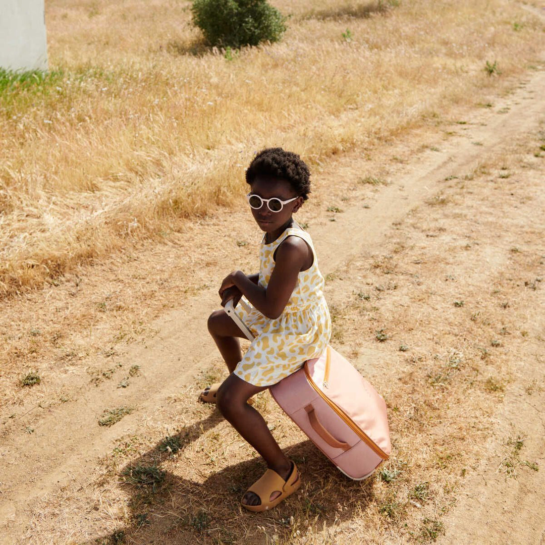 A girl wearing sunglasses and a dress is seating on a pink suitcase