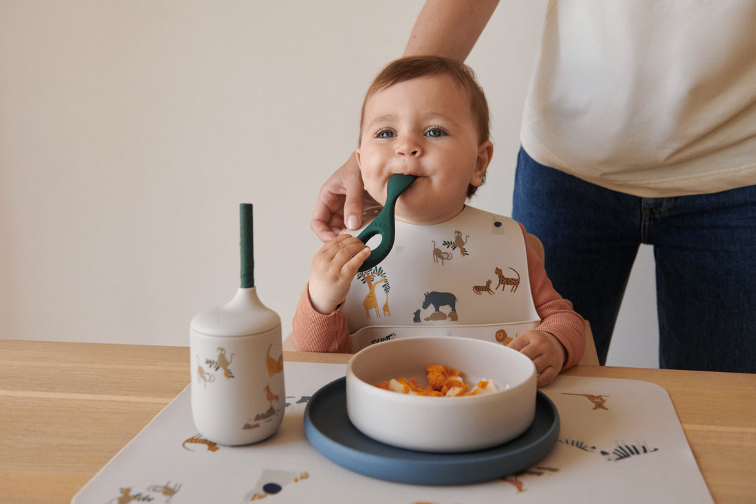 a toddler with a spoon in his mouth, wearing a bib with animal print and sitting at the table with a plate and drinking cup