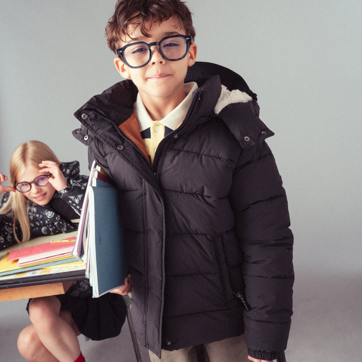 A young boy wearing thick black glasses and a black puffer jacket over a school uniform stands in front of a gray wall, holding a stack of books. A girl with glasses sits at a desk in the background.