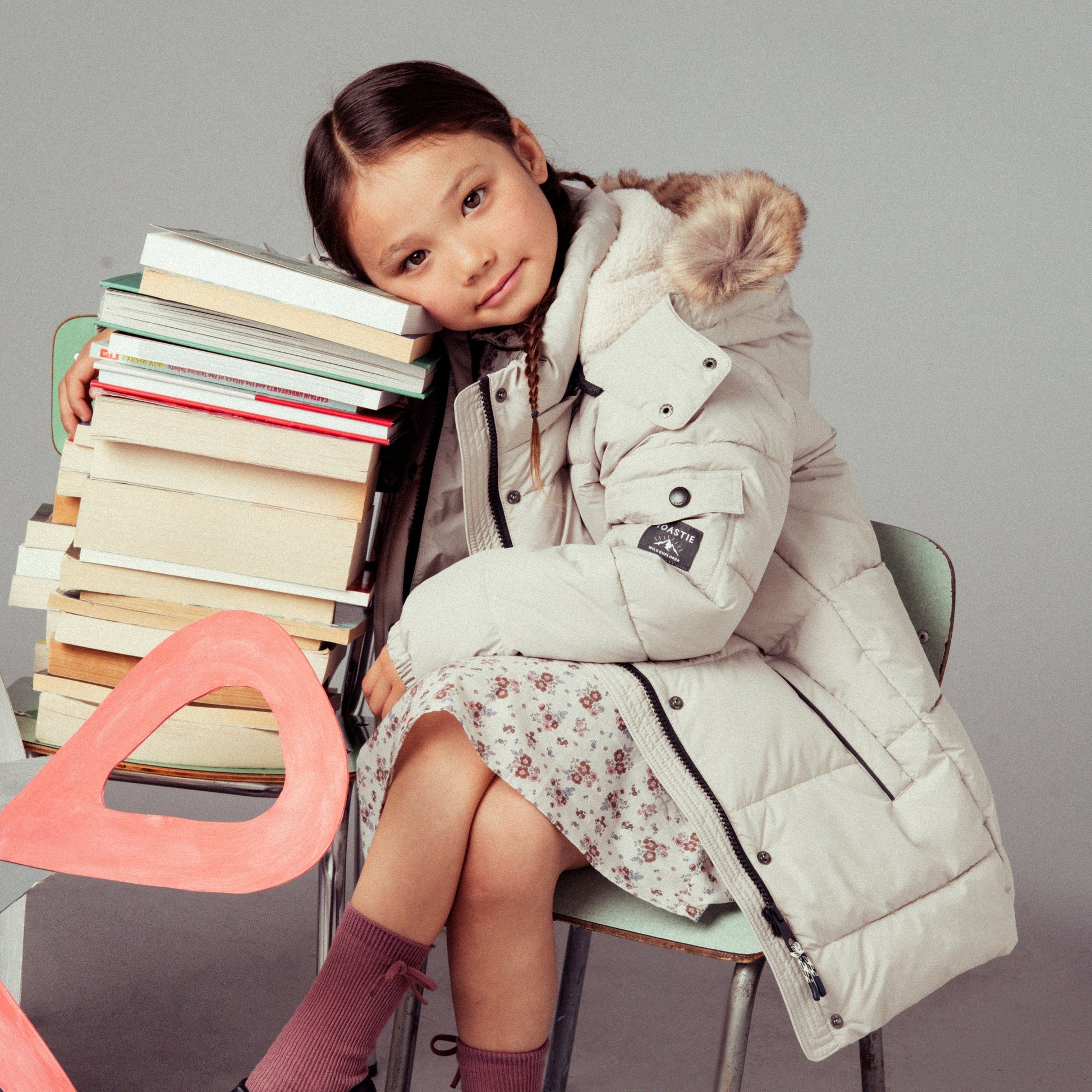 A young girl sits on a chair next to a stack of books, wearing a long cream puffer coat with a fur-trimmed hood, a floral skirt, and black Mary Jane shoes against a light gray studio background.