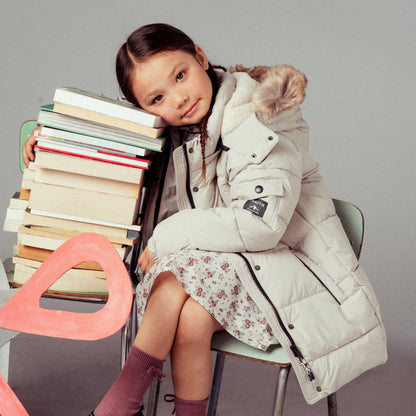 A young girl sits on a chair next to a stack of books, wearing a long cream puffer coat with a fur-trimmed hood, a floral skirt, and black Mary Jane shoes against a light gray studio background.