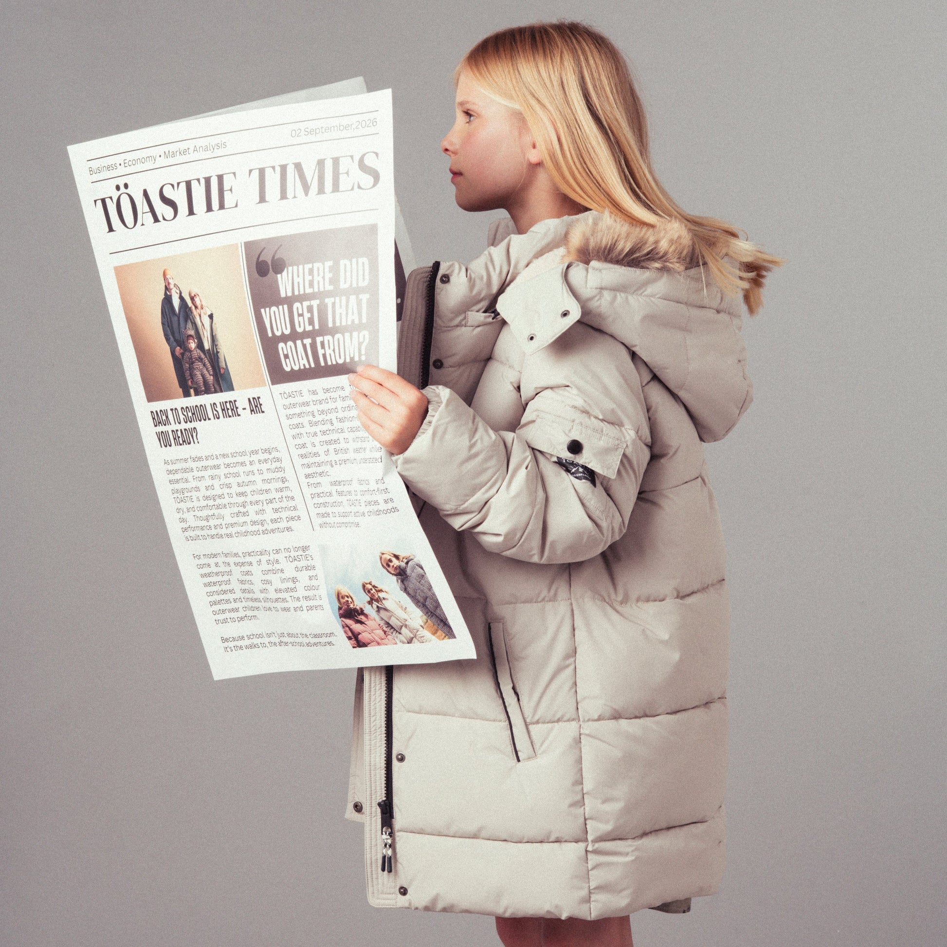 A young girl stands in profile wearing a long, light beige puffer coat and holding a oversized printed newspaper that reads 'The Toastie Times' against a plain gray studio background.