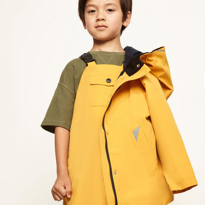 A young boy modeling a bright yellow hooded rain jacket over an olive green short-sleeve shirt, photographed against a clean off-white studio background.