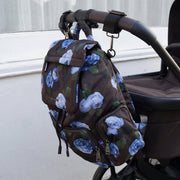 A brown backpack with a blue and white floral print hanging from the handle of a stroller outdoors next to a white wall.