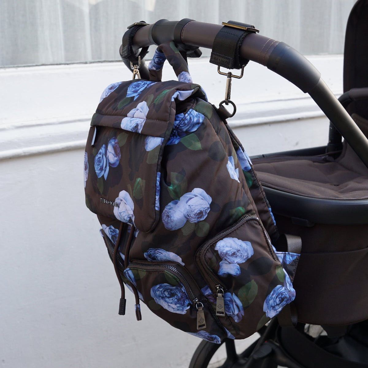 A brown backpack with a blue and white floral print hanging from the handle of a stroller outdoors next to a white wall.