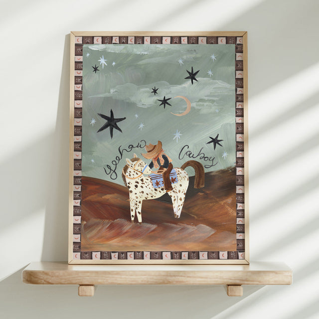 Framed artwork of a cowboy on a horse with stars and moon in the background, displayed on a shelf.