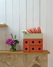 A styled interior scene showing the MODU Baby Walker in Burnt Orange which is a terracotta-colour. Designed in a storage box with circular cutouts and a cream-colored top tray holding round orange discs, placed on a wooden shelf beside a glass vase of purple flowers against a white paneled wall.