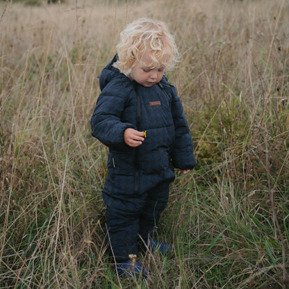 A toddler with curly blonde hair stands in a field of dry grass wearing a dark navy quilted snowsuit and matching pants.