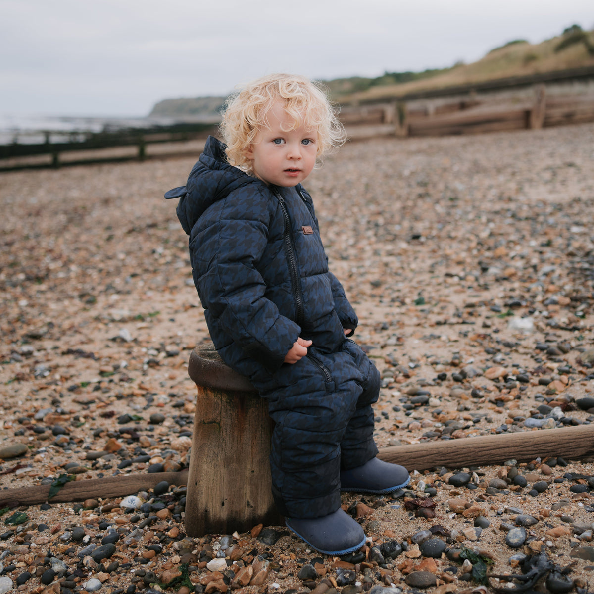 A toddler with curly blonde hair sits on a wooden stump on a pebble beach, wearing a dark navy quilted puffer snowsuit with welly boots.