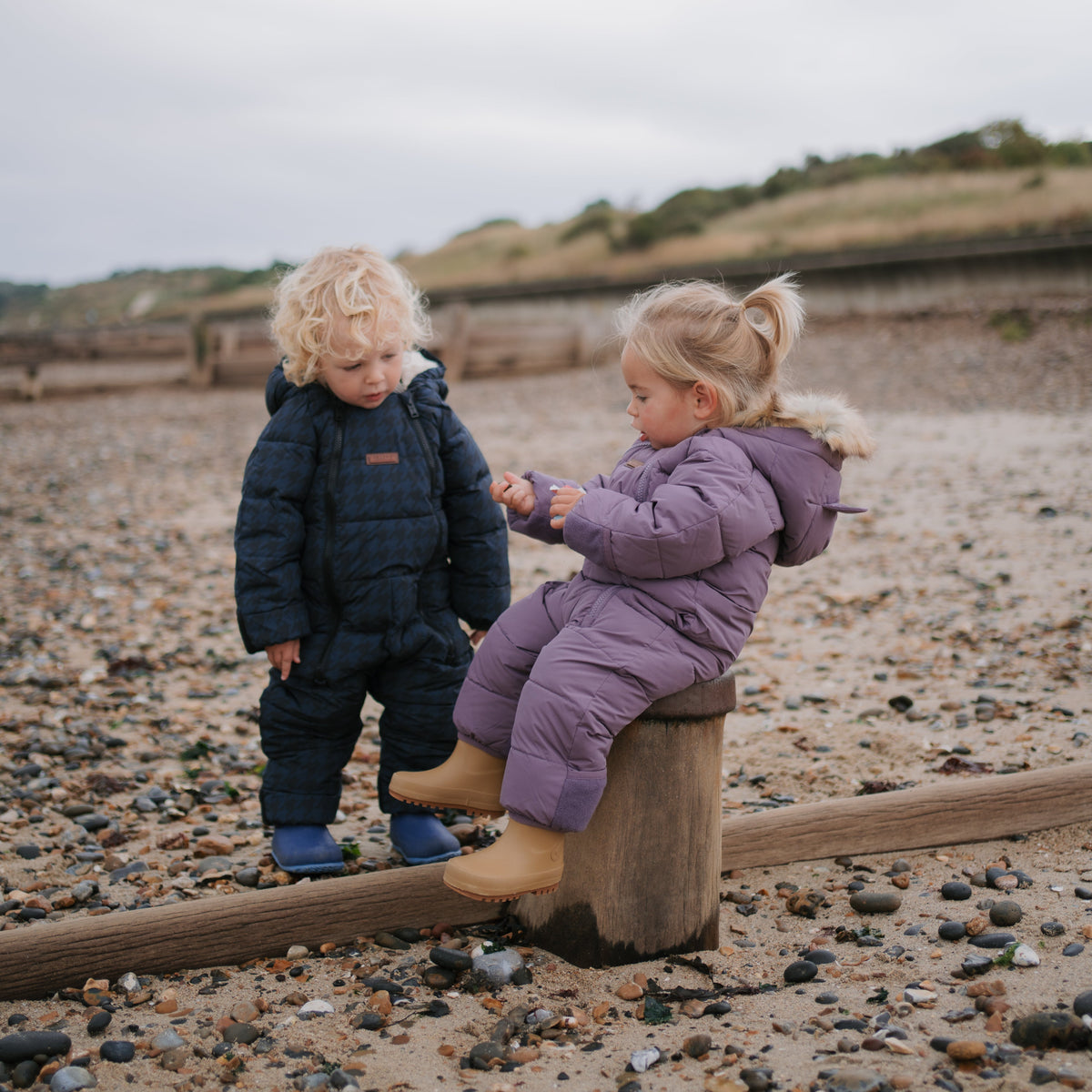 Two young children in winter clothing on a pebble beach. One child stands wearing a dark navy quilted snowsuit, and the other sits on a wooden post wearing a lavender quilted snowsuit and tan boots.