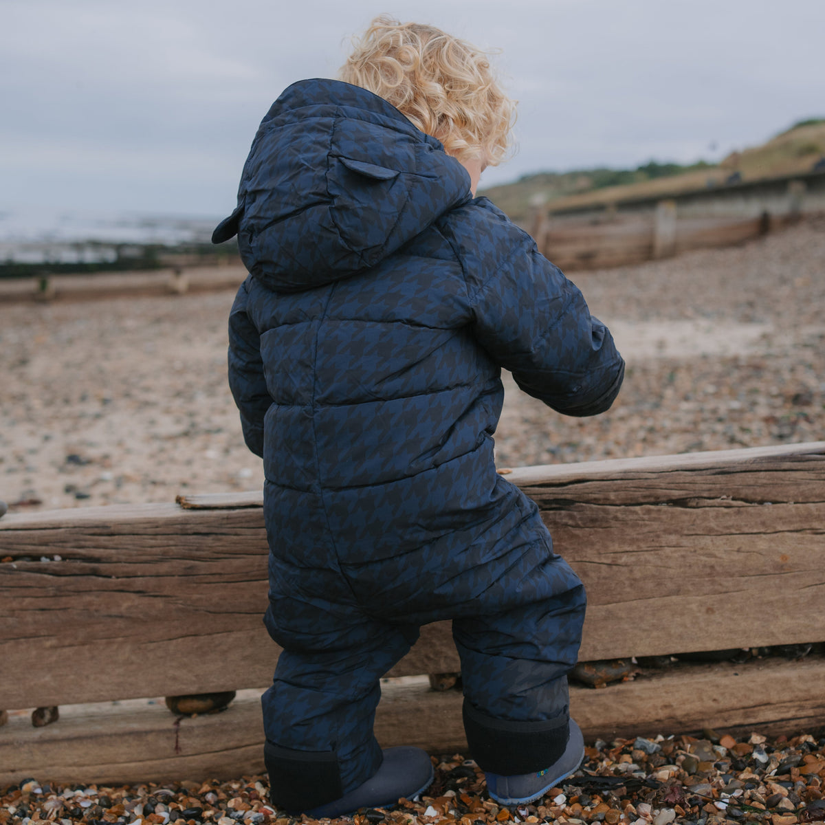 A toddler with curly blonde hair seen from behind, wearing a dark navy quilted snowsuit and standing on a pebble beach next to a wooden fence under an overcast sky.
