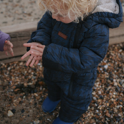 A toddler with curly blonde hair wearing a navy blue puffer snowsuit and blue rain boots, standing on a pebble beach.
