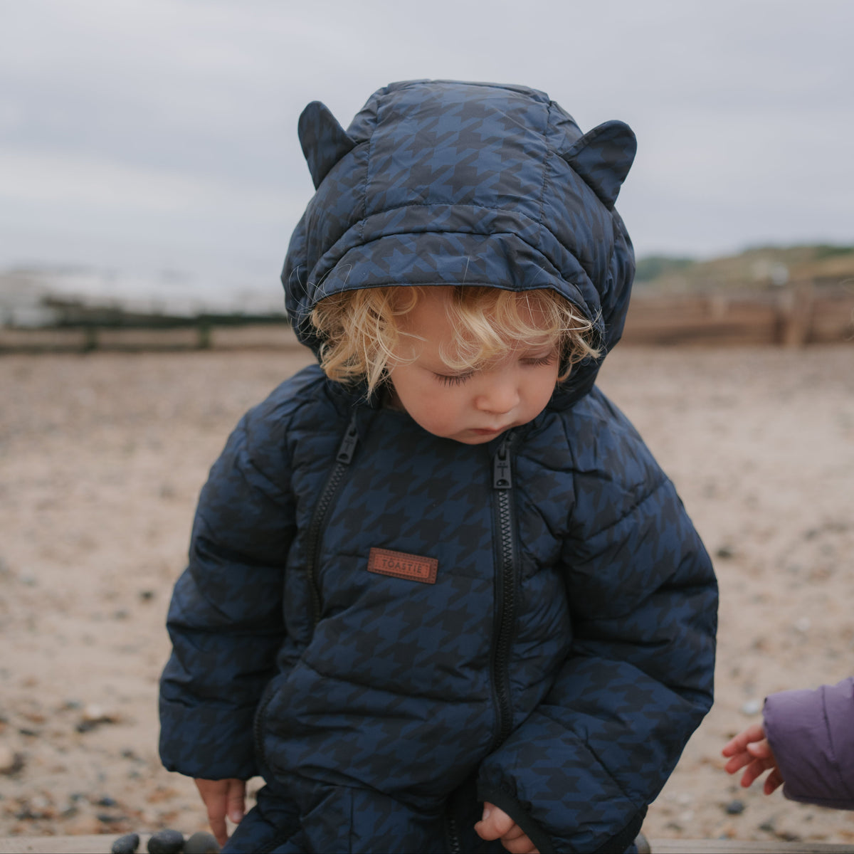 A toddler wearing a navy blue patterned puffer snowsuit with a bear-ear hood, standing on a sandy beach under an overcast sky.