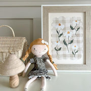 Doll with red hair and a flower in her hair next to a framed artwork of flowers on a light-colored surface.
