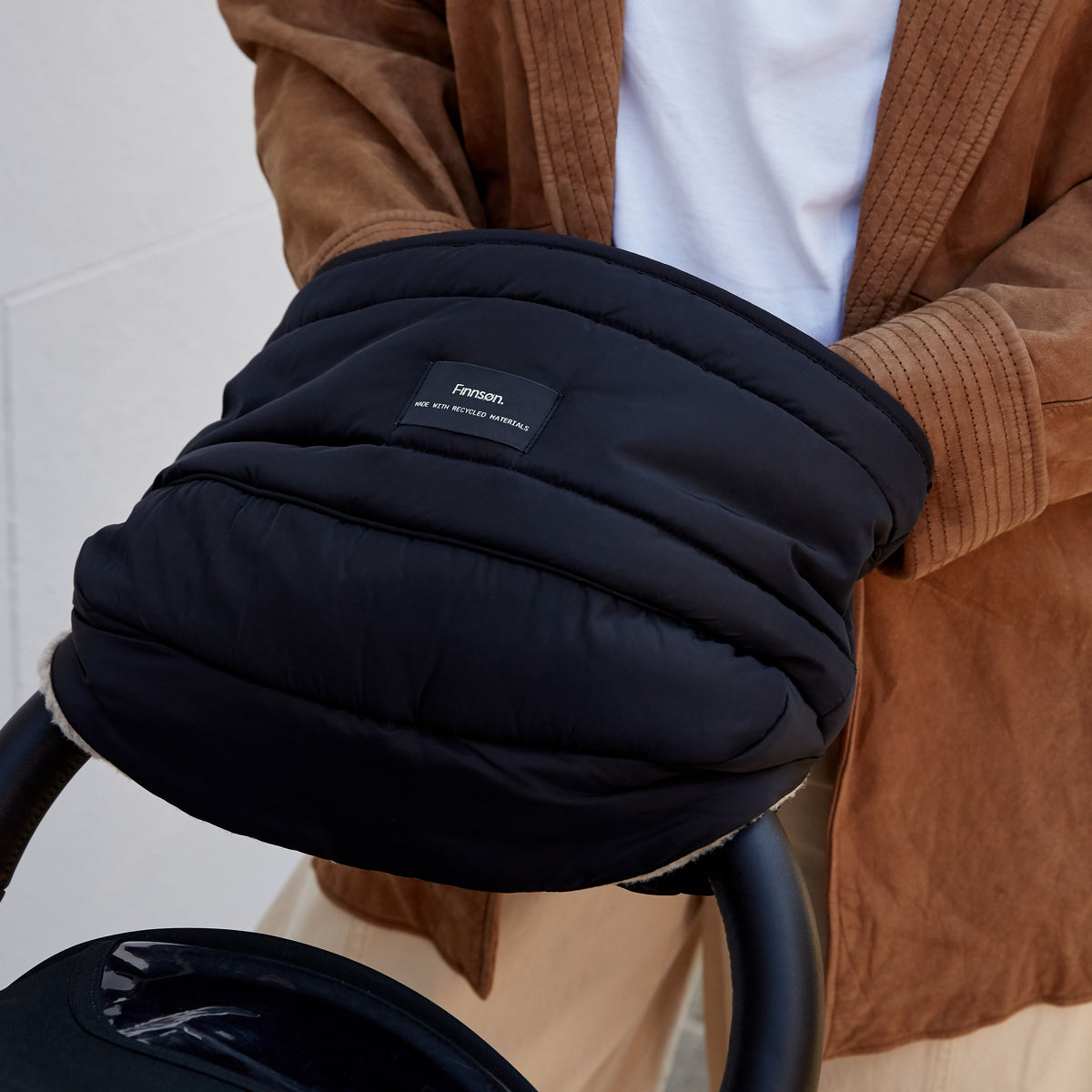 A close-up of a person wearing a Finnson black quilted puffer mono mitten pushing a black pushchair stroller. 