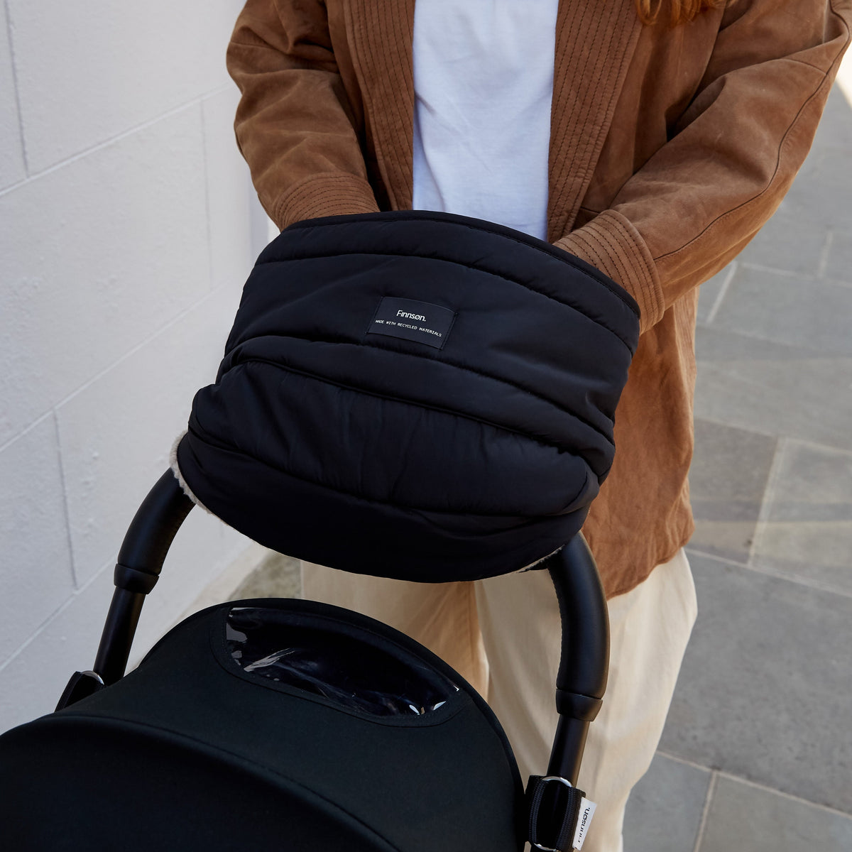 A person wearing a brown corduroy jacket pushing a black stroller with a Finnson padded black Mono Mitten featuring a small white brand label that reads 'finnson'. The setting is outdoors against a light grey stone wall.