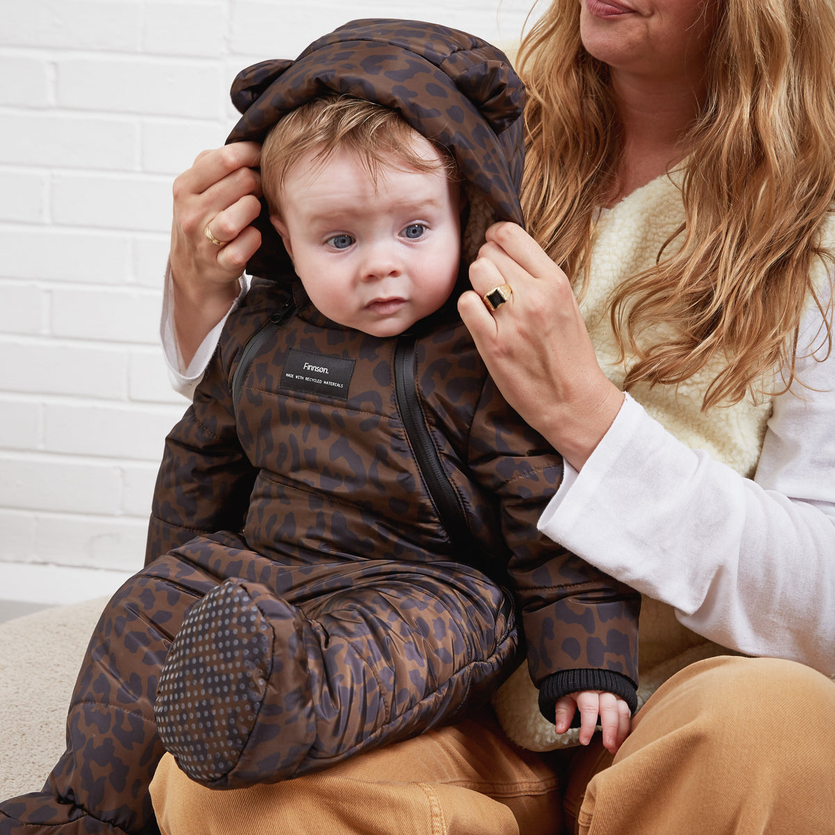 A baby wearing a brown and black leopard-print footed sleeper with a bear-ear hood, sitting on a woman's lap against a white brick wall.