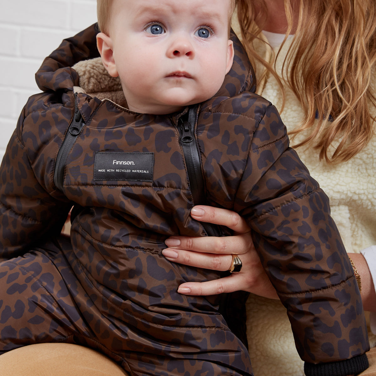 A baby wearing a Finnson brown leopard-print hooded snowsuit, held by a woman against a white brick wall background.