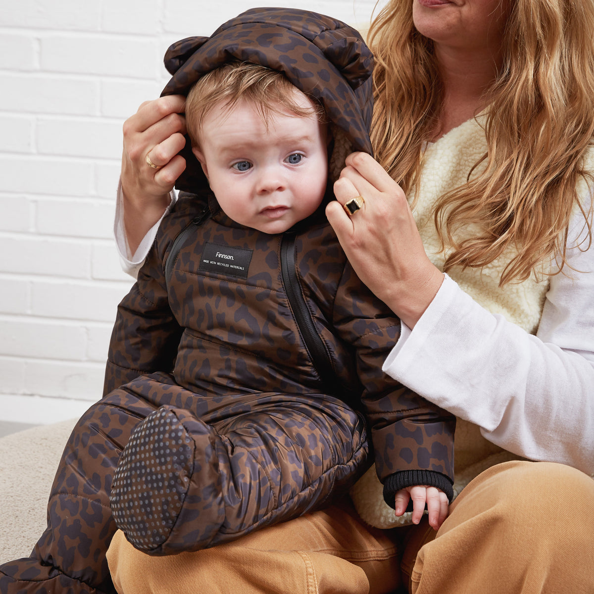 A baby wearing a Finnson brown and black leopard-print snowsuit with a footed sleeper, bear-ear hood, sitting on a woman's lap against a white brick wall.
