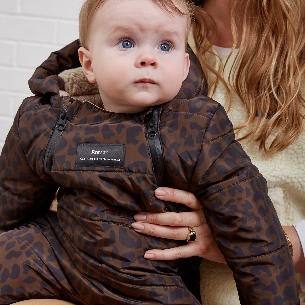 A baby wearing the Finnson brown leopard-print hooded snowsuit, held by a woman against a white brick wall background.