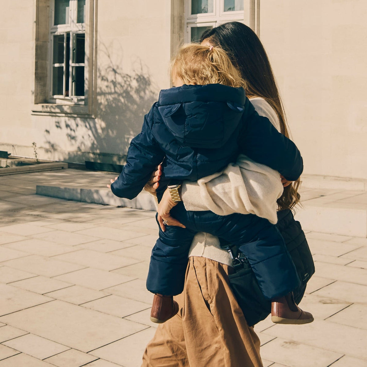 A woman carrying a child outdoors, wearing the Finnson dark navy puffer Snowsuit. The woman also wears a cream-colored top and tan trousers.