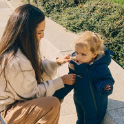 A woman crouches outdoors next to a child wearing a Finnson navy blue hooded winter snowsuit, while the woman adjusts the jacket. The setting includes a concrete walkway and green shrubs in the background.