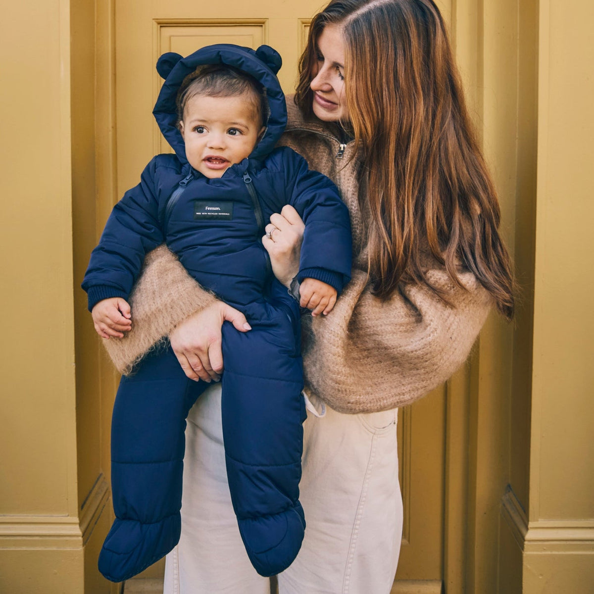 A woman holds a baby wearing a Finnson navy blue puffer snowsuit with bear ears on the hood. The baby is standing in front of a warm yellow door.