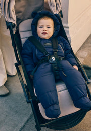 A baby wearing a Finnson navy blue puffer snowsuit with a hooded bear-ear design, sitting in a stroller.