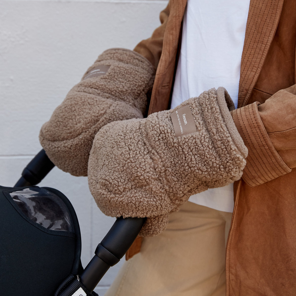 A close-up of a person wearing Finnson cozy taupe fleece winter stroller mittens in a brown corduroy jacket, and a white t-shirt, holding a black stroller handle against a white brick wall.