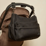 Finnson black diaper bag with a front zippered pocket and side gusset, shown hanging from the handle of a brown stroller against a plain beige wall.