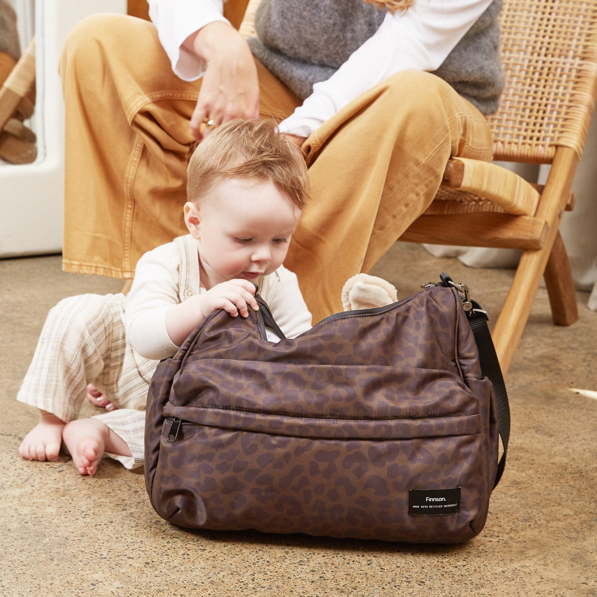 A woman and a baby sit on a carpeted floor in a warm, neutral-toned room, with a Finnson dark brown patterned changing diaper bag in the foreground. 