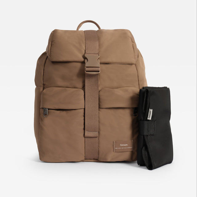 A tan Finnson Changing backpack with a central buckle strap, shown next to a black rolled travel changing mat on a light gray background. The backpack features a top flap, front zip pocket, and a small tan brand patch near the bottom right.