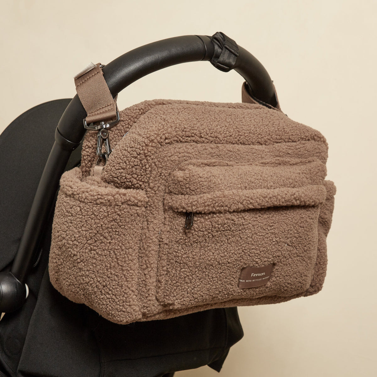 Finnson mocha brown borg oversized stroller organiser bag with stroller clips and a small brown brand patch, shown hanging from a black stroller handle against a plain beige background.