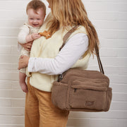 A woman holding a baby stands against a white brick wall, wearing a mocha brown borg Finnson shoulder bag across her body. The woman wears a cream vest over a white long-sleeve shirt and tan pants.