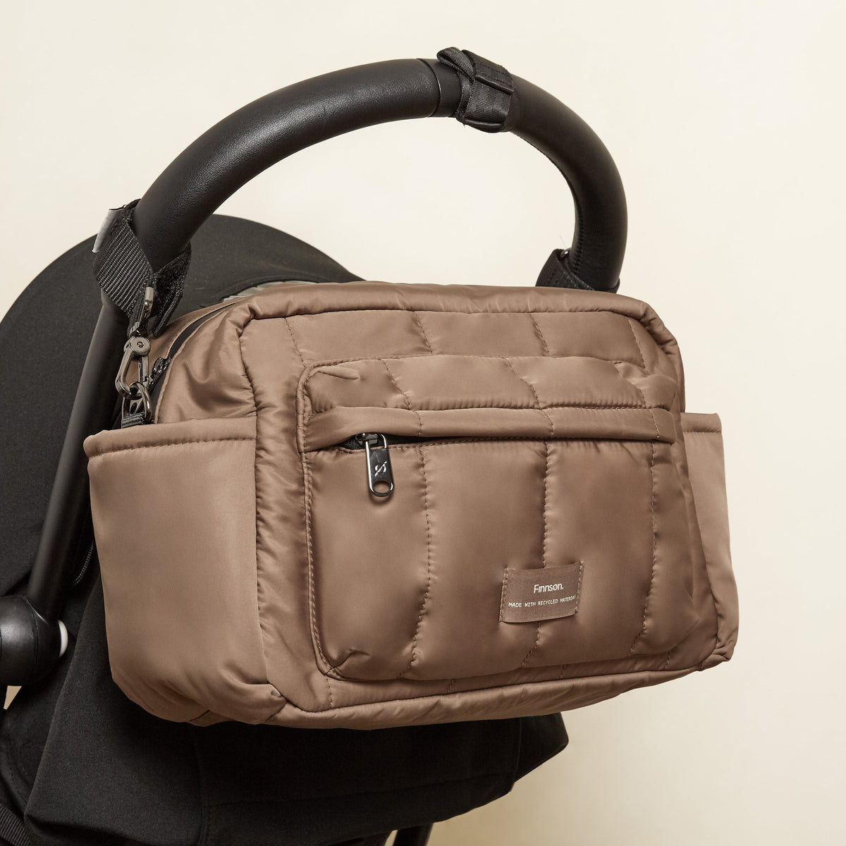A Finnson mocha brown oversized stroller organiser bag with a front zippered pocket, shown hanging from a black stroller handle.
