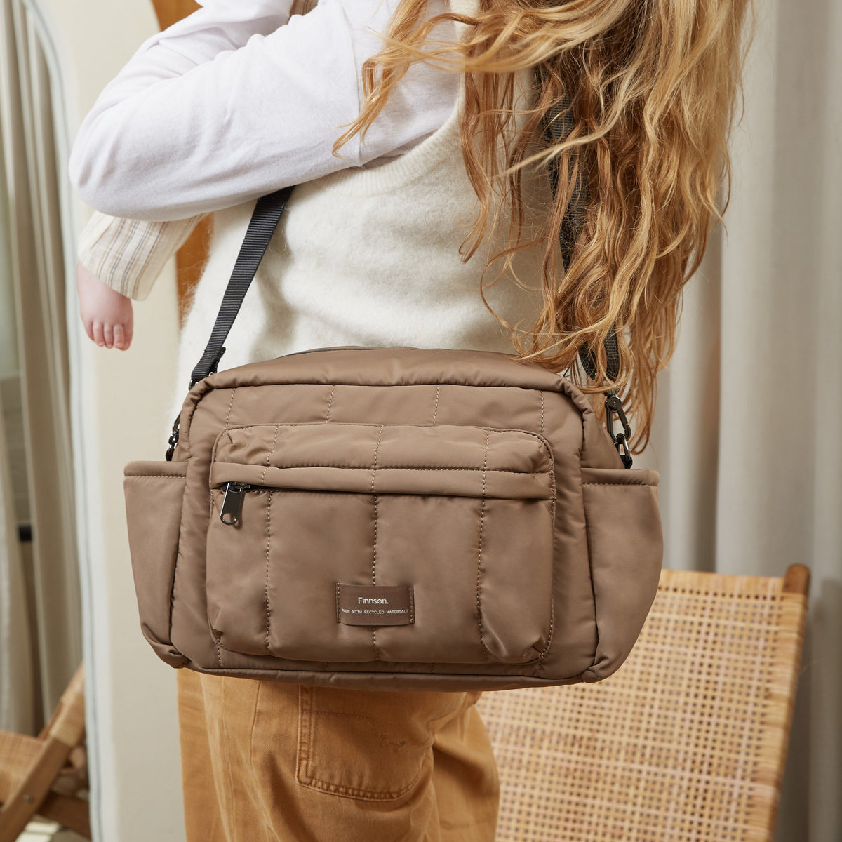 A woman carries a mocha brown Finnson crossbody oversized stroller organiser bag with a black strap in an indoor setting.