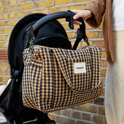A person holds a black stroller next to a tan and black gingham diaper bag with a white 'Fenian' label, set against a yellow brick wall.