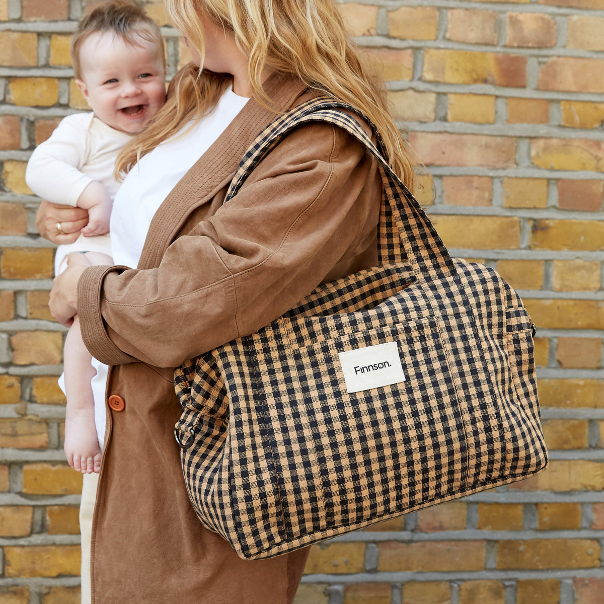 A woman carrying a baby on her back while wearing a brown corduroy jacket and carrying a large brown and black gingham check shoulder bag with a white 'Ferson' label, set against a brick wall background.