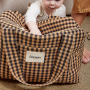 A toddler sits inside a large brown and black gingham check bag, with an adult's hand and legs visible in the background. The bag features a front pocket with a white label that reads 'Finnson'.