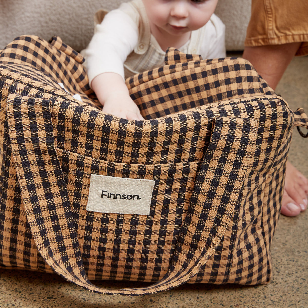 A toddler sits inside a large brown and black gingham check bag, with an adult's hand and legs visible in the background. The bag features a front pocket with a white label that reads 'Finnson'.