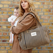 A woman in a brown coat holding a baby, carrying a large brown and black checkered duffel bag over her shoulder, set against a yellow brick wall.