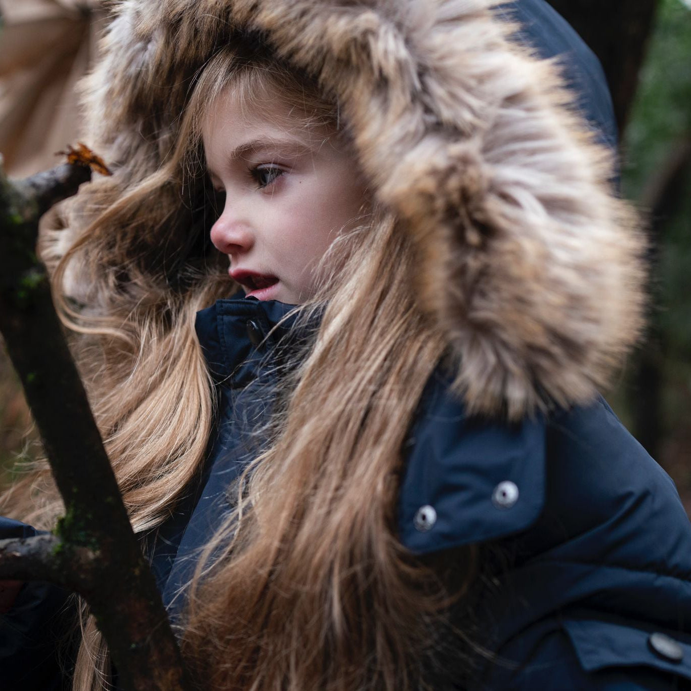 A young girl wearing a dark navy winter parka with a large brown faux-fur trimmed hood, holding a small branch in a wooded outdoor setting.