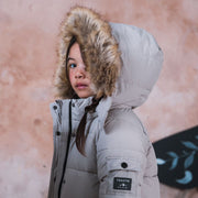 A child wearing a light gray puffer jacket with a brown faux-fur trimmed hood, standing against a textured pinkish-beige wall. The jacket has a black brand patch on the sleeve that reads 'TOASTIE'.