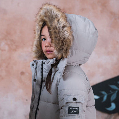 A child wearing a light gray puffer jacket with a brown faux-fur trimmed hood, standing against a textured pinkish-beige wall. The jacket has a black brand patch on the sleeve that reads 'TOASTIE'.