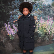 A child wearing a long navy blue puffer parka with a faux-fur trimmed hood, standing in front of a painted forest backdrop.