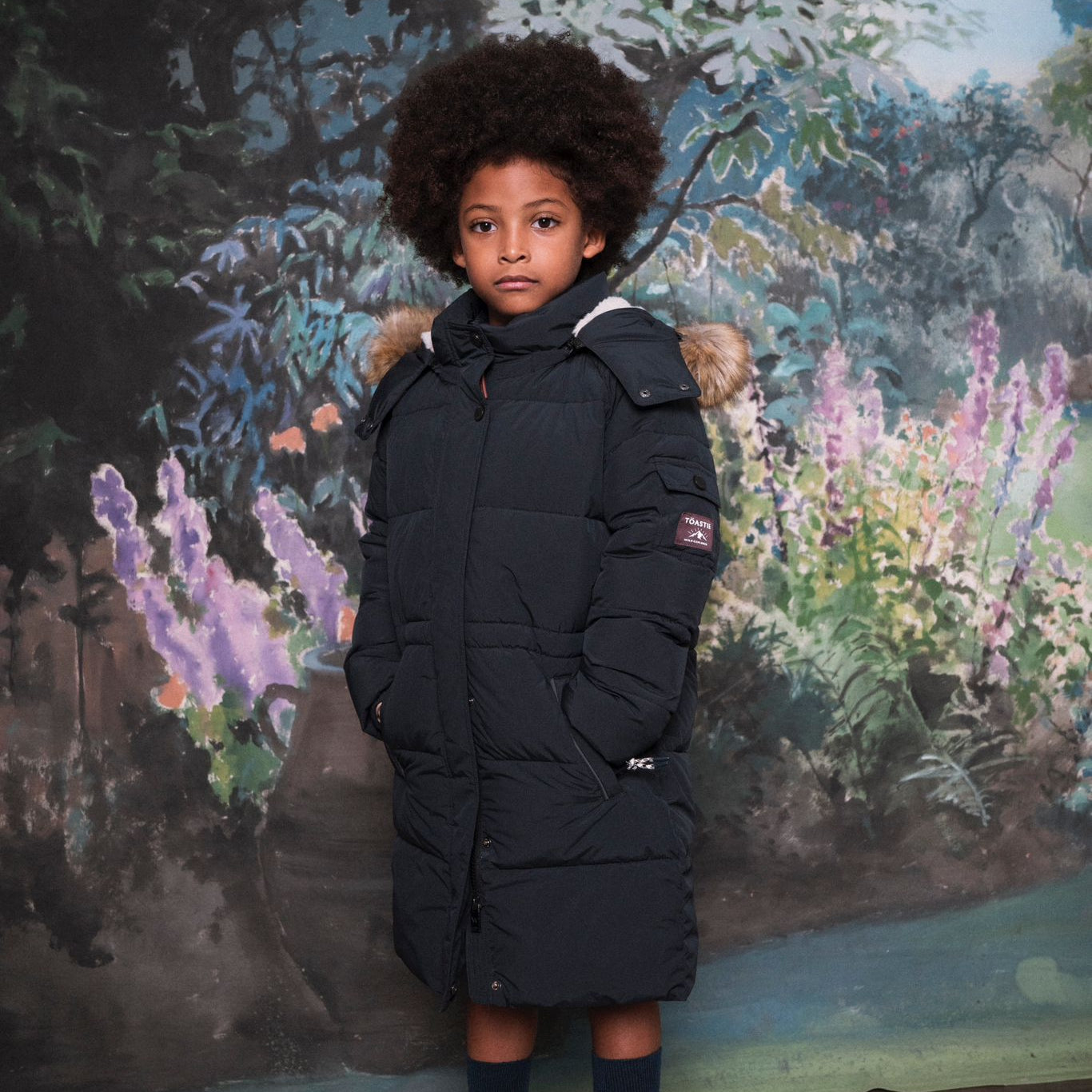 A child wearing a long navy blue puffer parka with a faux-fur trimmed hood, standing in front of a painted forest backdrop.