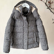 A black and white houndstooth patterned puffer jacket with a beige sherpa-lined hood, hanging on a wooden hanger labeled 'TOASTIE' against a beige wall with a decorative branch in the background.