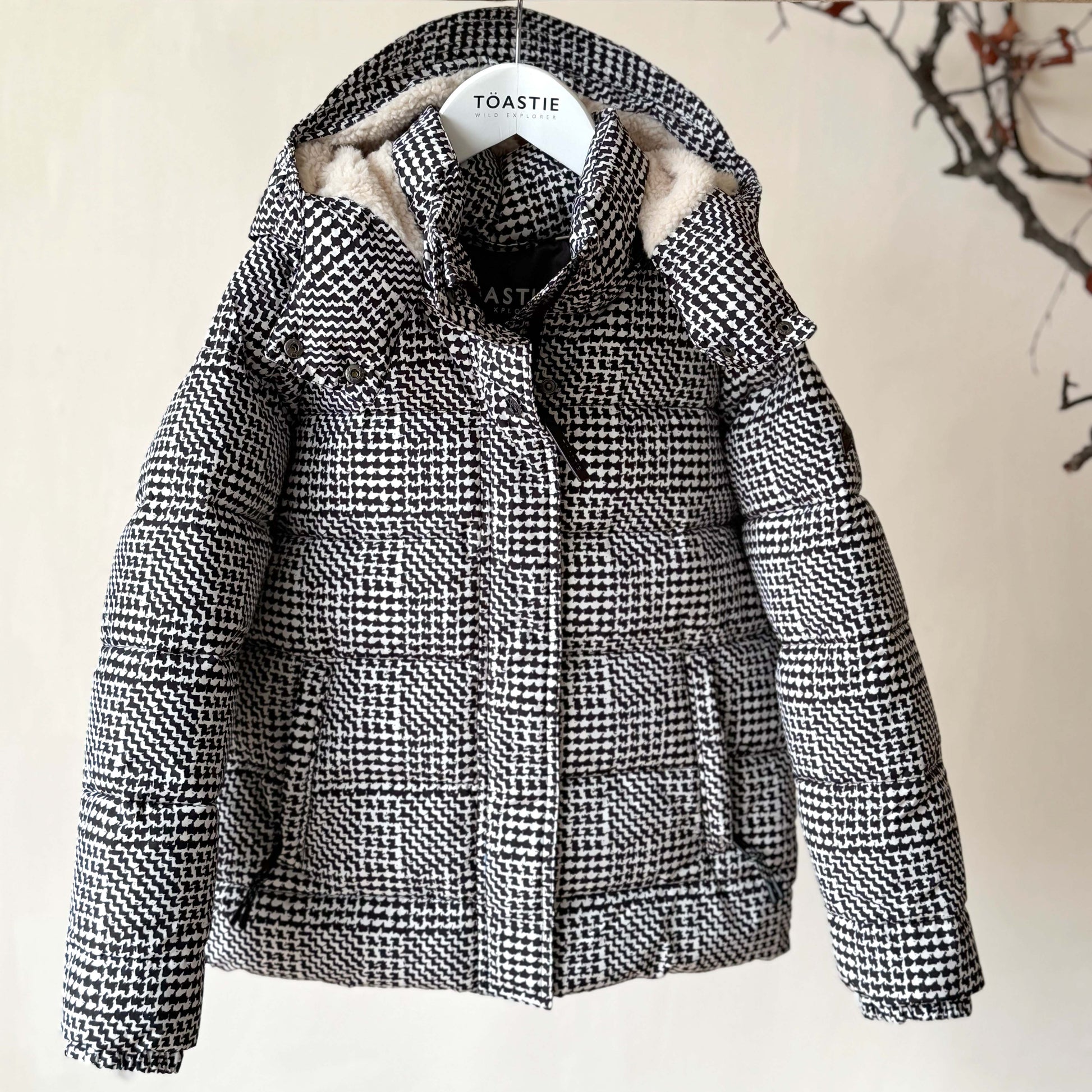 A black and white houndstooth patterned puffer jacket with a beige sherpa-lined hood, hanging on a wooden hanger labeled 'TOASTIE' against a beige wall with a decorative branch in the background.