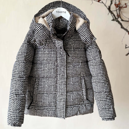 A black and white houndstooth patterned puffer jacket with a beige sherpa-lined hood, hanging on a wooden hanger labeled 'TOASTIE' against a beige wall with a decorative branch in the background.