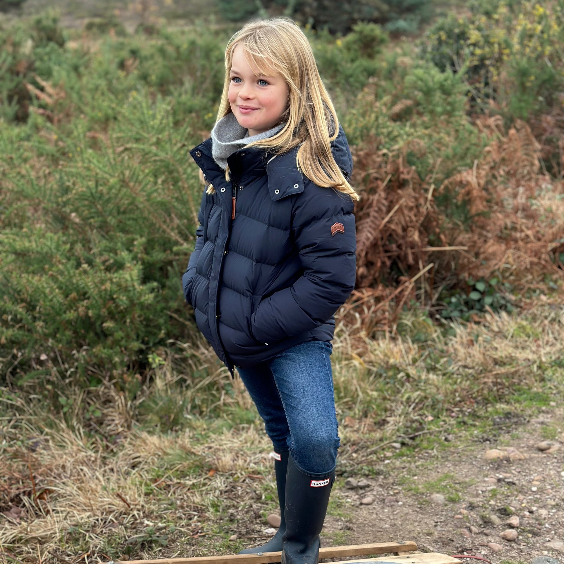 A young girl stands on a wooden sled outdoors in a grassy, wooded setting. She is wearing a navy blue puffer jacket, a gray knit scarf, blue jeans, and black rubber boots.
